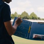 Football coach with blue board for tactics stood next to pitch