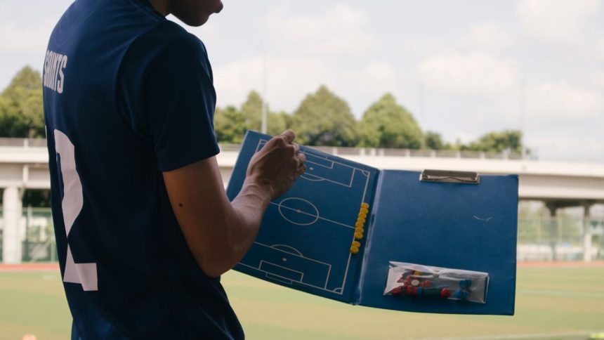 Football coach with blue board for tactics stood next to pitch