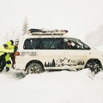 Two people push a stuck van in the snow.