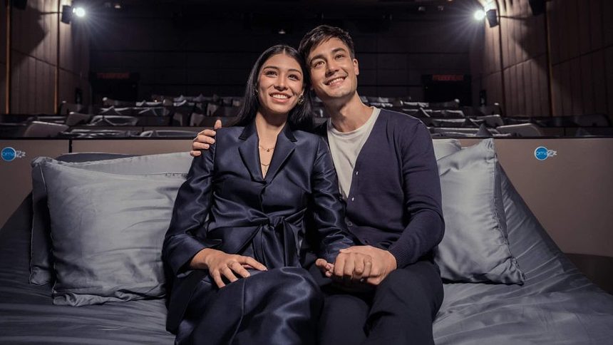 A promotional image of a couple sitting on an Omazz bed in 'The Bed Cinema.'