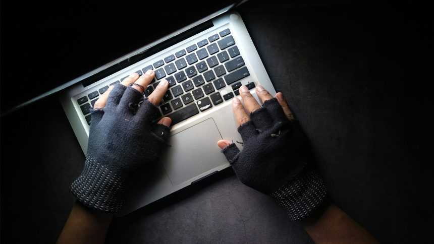 A man with gloved hands uses a laptop in the dark.